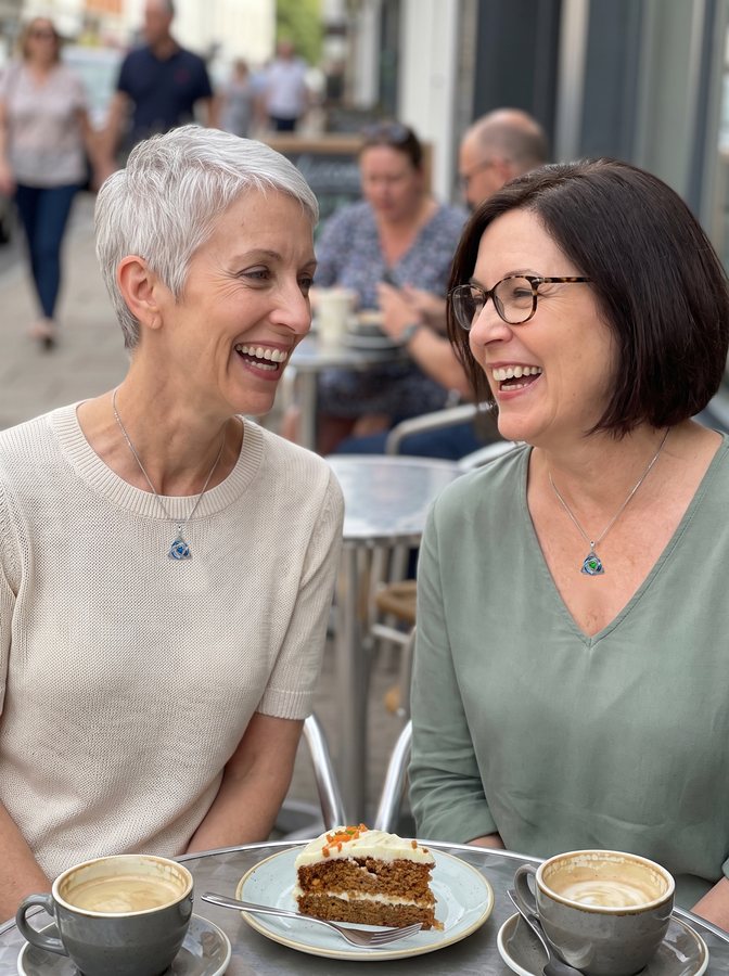 Two friends laughing on a café terrace, each wearing her necklace